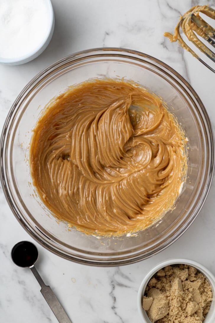 peanut butter and butter mixed together in a glass bowl on a white marble countertop with other ingredients around it.
