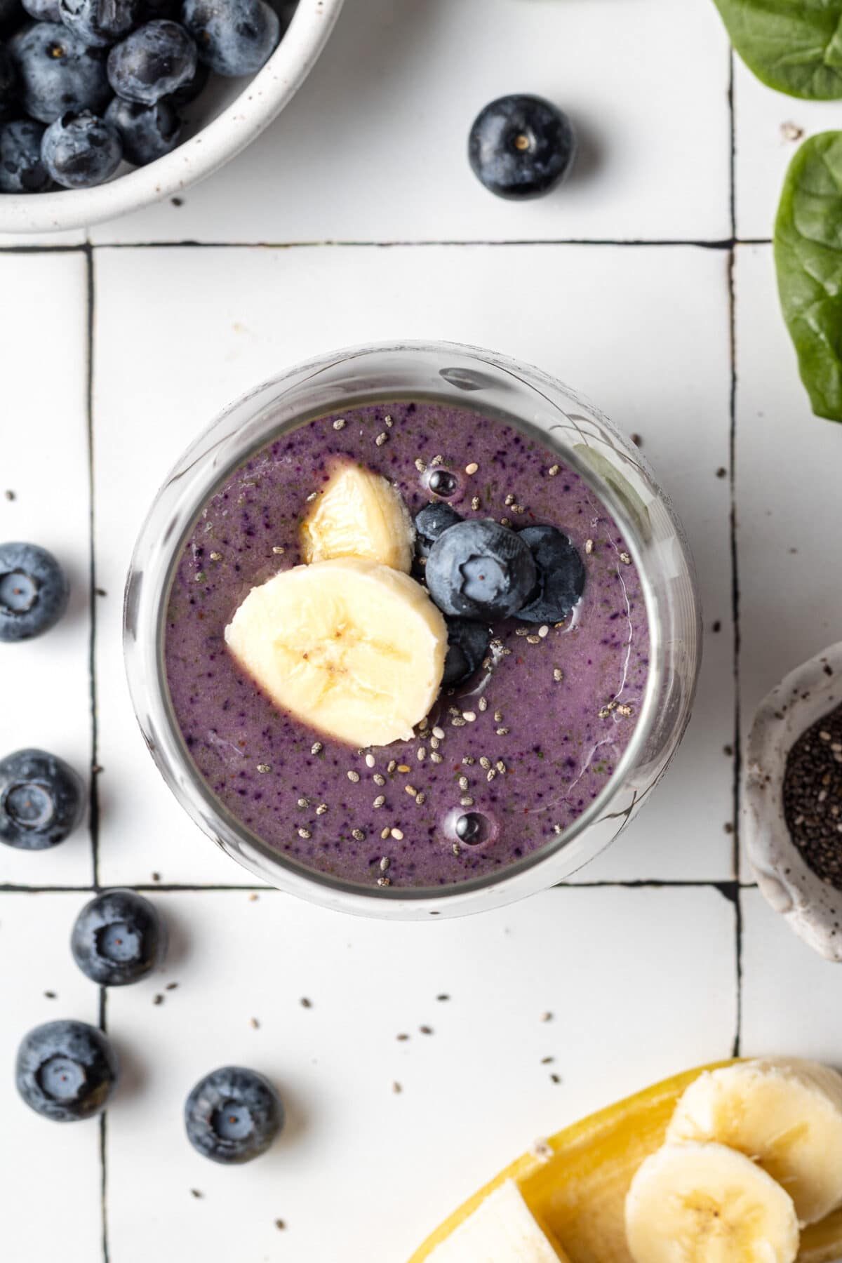 blueberry banana spinach smoothie in a glass with bananas and blueberries scattered around and chia seeds on top