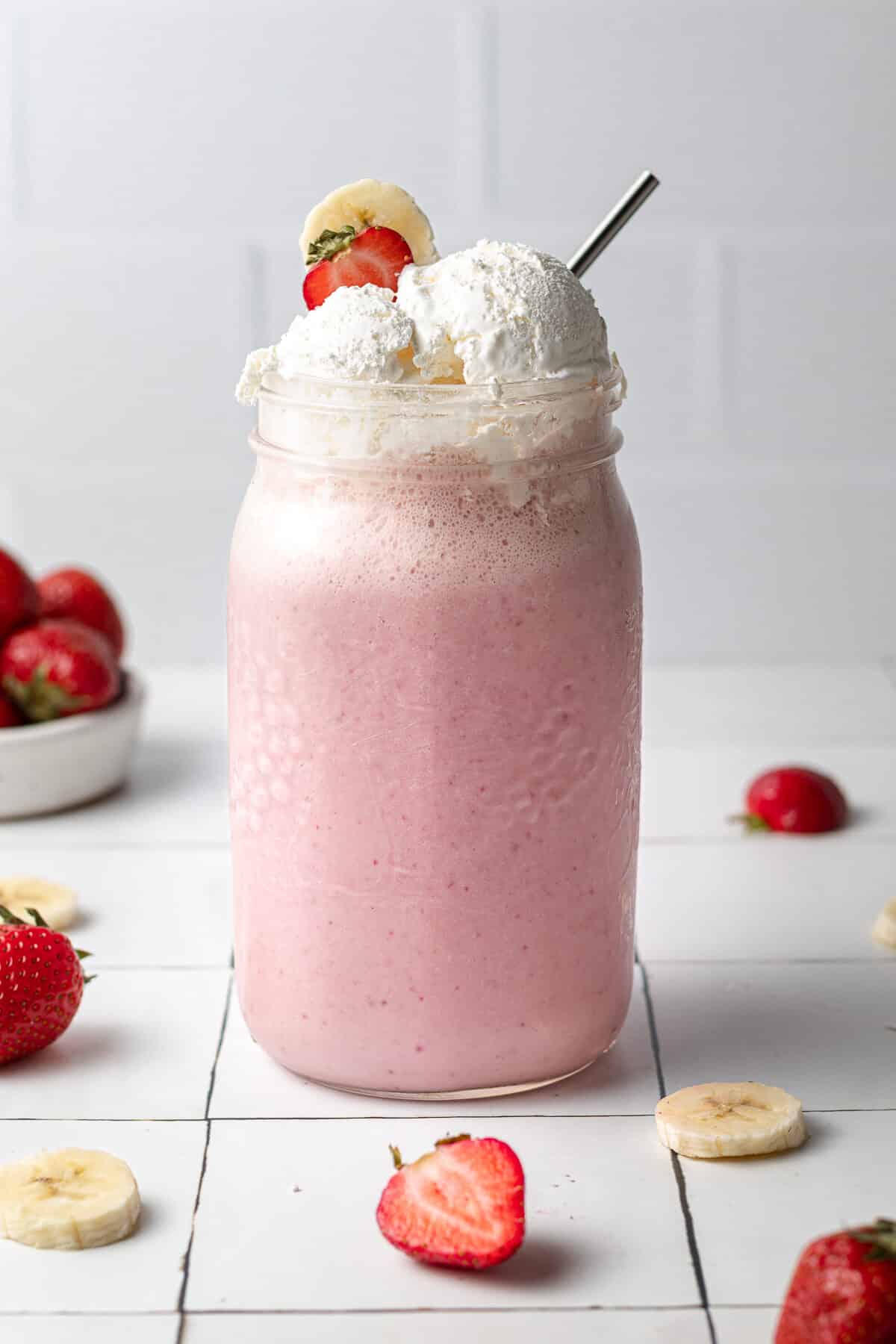 strawberry banana milkshake in a large glass with whipped cream on top and fruit around it
