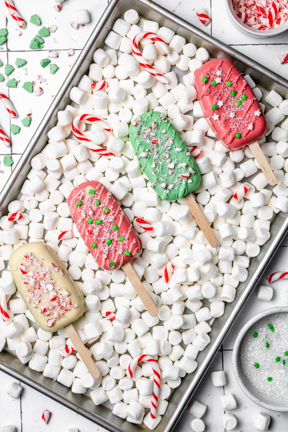 christmas cakesicles on a baking tray with marshmallows and candy canes around them