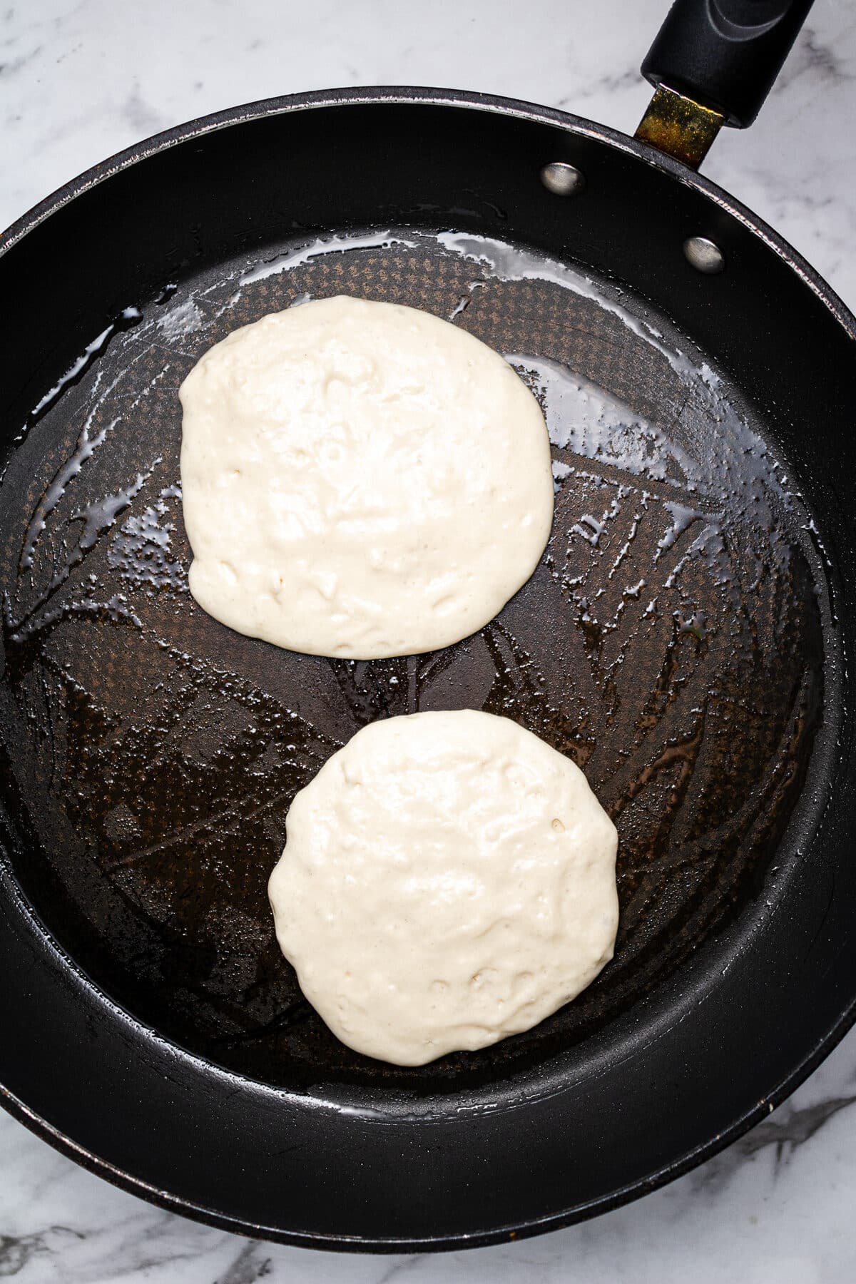 Two pancakes cooking on a black frying pan.