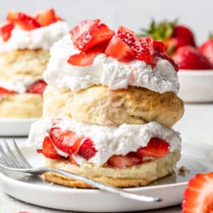 Vegan strawberry shortcake pictured on a plate with a bowl of strawberries and another shortcake cake in the background