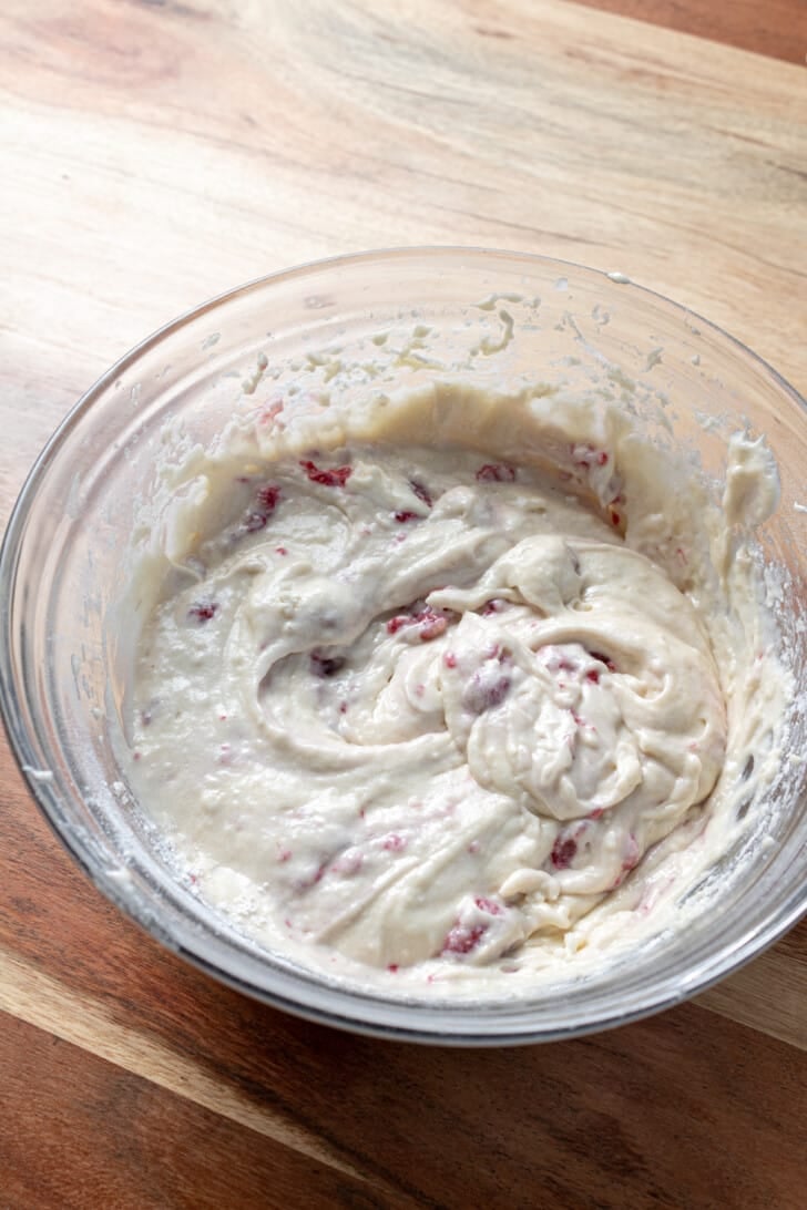 cupcake batter with raspberries mixed into it in a glass mixing bowl on a wooden countertop.