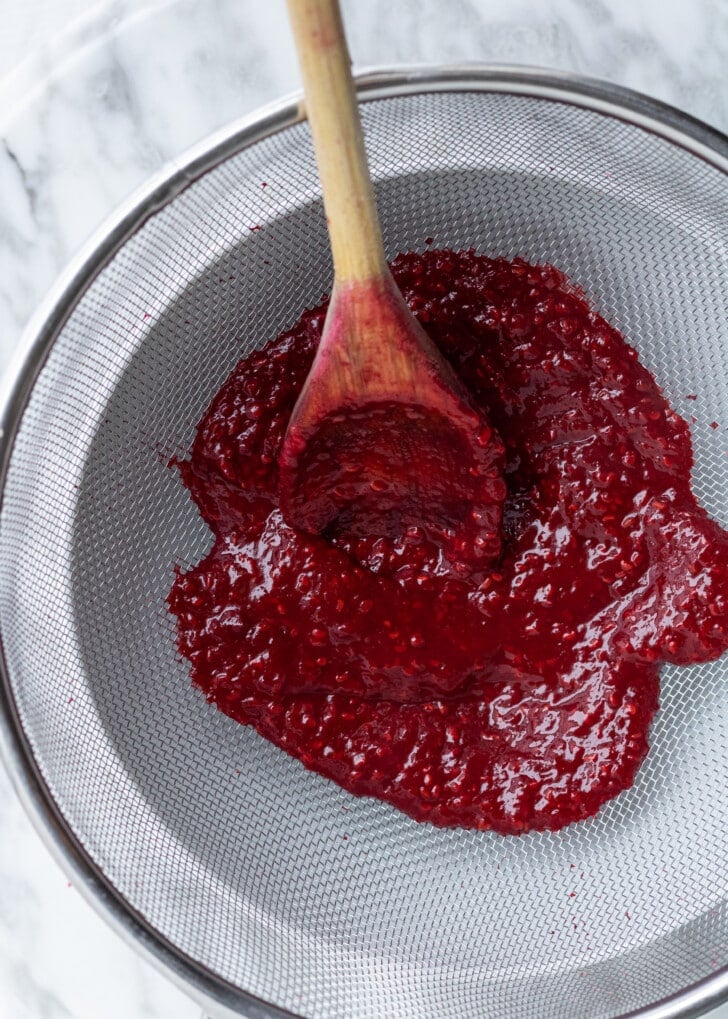 a mesh sieve over a white bowl with a wooden spoon inside squishing raspberry puree into the bowl.