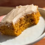 close up of a pumpkin bar on a white plate with frosting on top and a hand holding the plate.