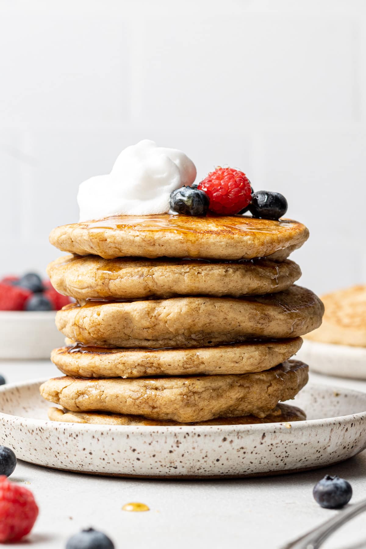 Tall stack of oat flour pancakes on a plate and topped with fresh whip cream and berries