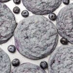 blue cookies on a baking sheet with blueberries scattered around them.