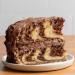 A slice of marble cake with swirled chocolate and vanilla layers is topped with rich chocolate frosting, set on a white plate against a wooden table.
