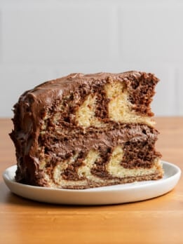 A slice of marble cake with swirled chocolate and vanilla layers is topped with rich chocolate frosting, set on a white plate against a wooden table.