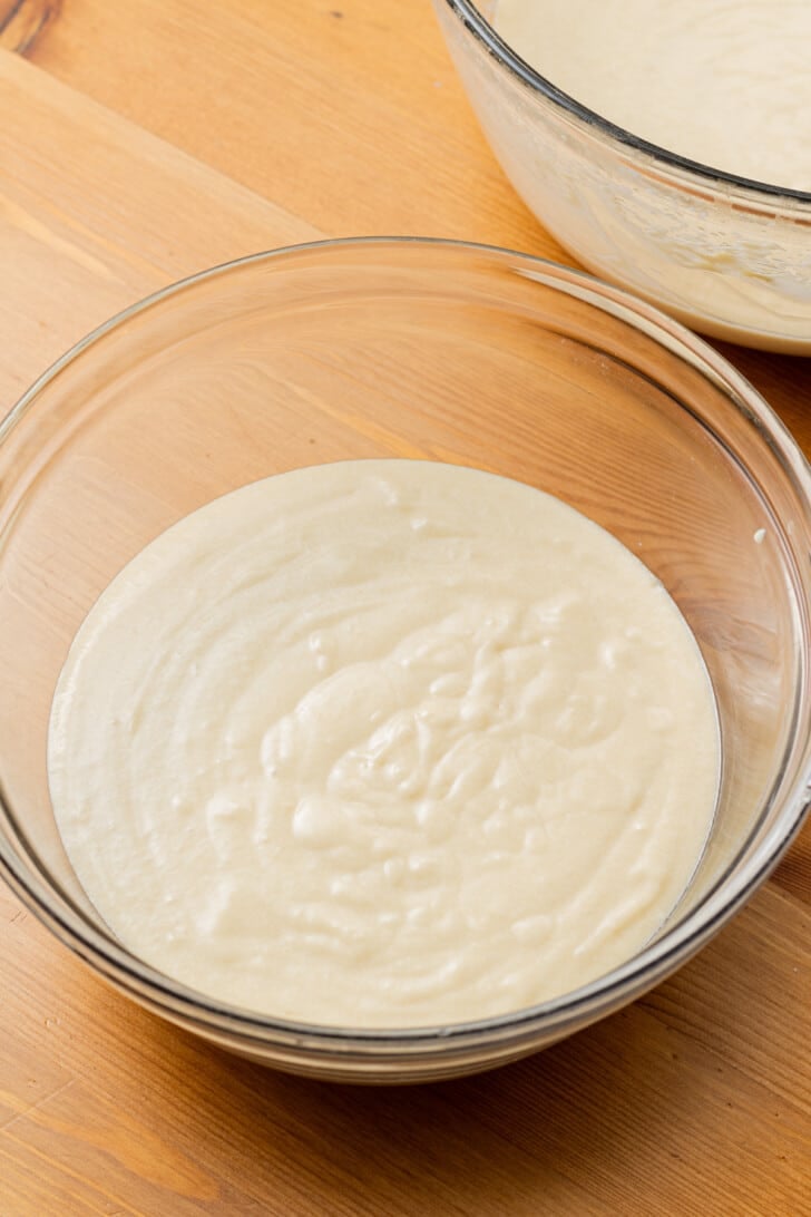 A glass bowl filled with smooth, creamy batter sits on a wooden table. Another bowl is partially visible in the background, suggesting baking preparations.