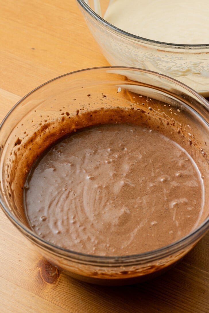 A glass bowl filled with smooth, creamy chocolate batter sits on a wooden table. Another bowl with light batter is partially visible in the background.
