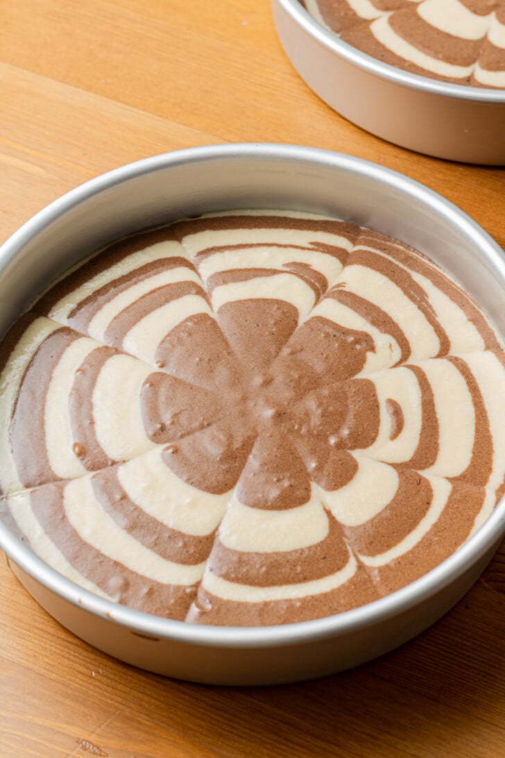A round pan filled with unbaked marbled cake batter, featuring alternating concentric circles of chocolate and vanilla, on a wooden surface.