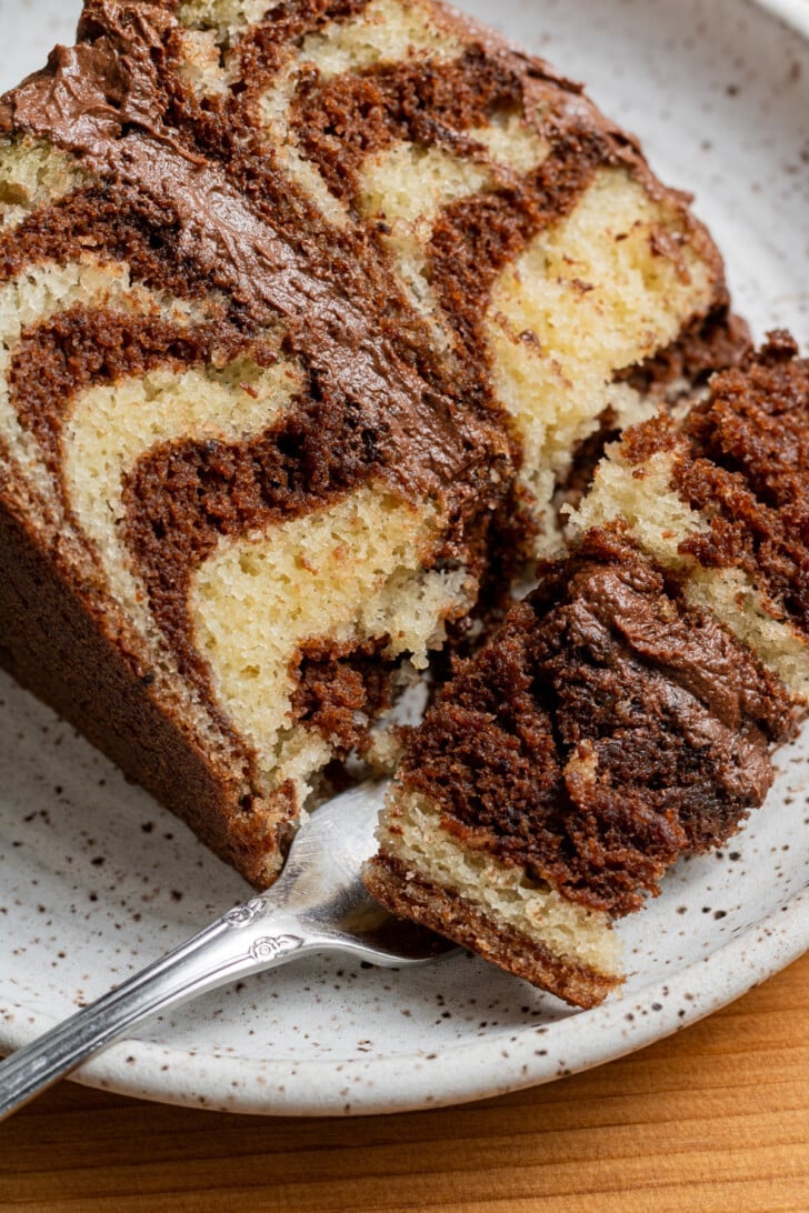 A close-up of a sliced marble cake with chocolate and vanilla swirls, on a speckled plate. A metal fork rests next to the cake slice. Warm and inviting.