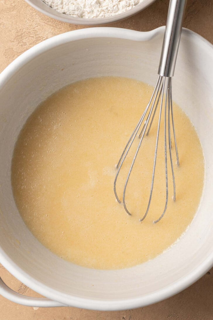 A metal whisk in a white bowl of smooth batter, with a bowl of flour in the background. The scene conveys a homey baking atmosphere.