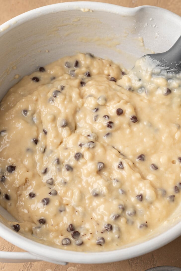 Bowl of thick banana chocolate chip muffin batter with visible chocolate chips. A spatula rests inside. The tone is warm and inviting.