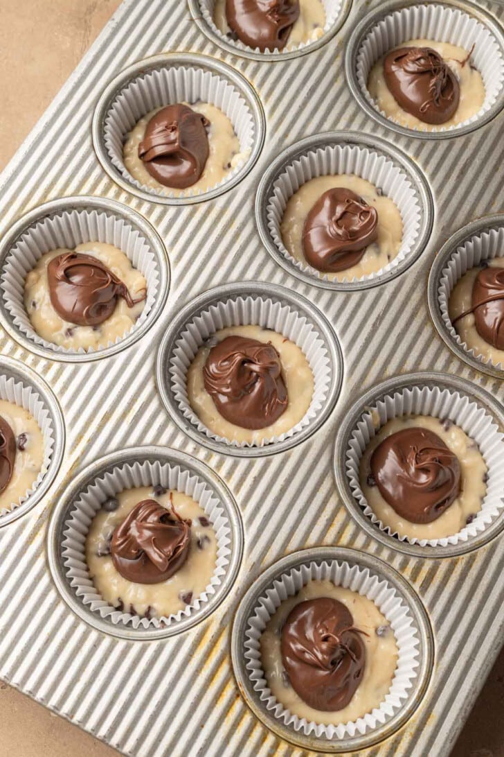 Cupcake tray filled with raw batter in paper liners, topped with dollops of chocolate spread. The image conveys a mood of anticipation and indulgence.