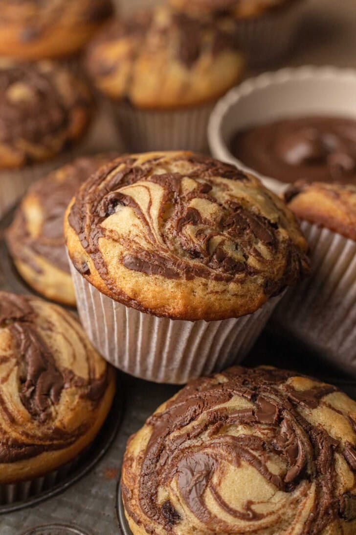 Close-up of swirled chocolate banana muffins in paper liners, showcasing a rich marbled pattern. Background shows blurred muffins and a bowl of chocolate spread. Cozy, inviting feel.
