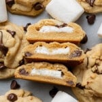 Close-up of chocolate chip cookies with melted marshmallow centers. Some are split open to reveal filling, surrounded by chocolate chips and marshmallows. Cozy and delicious.