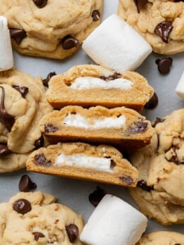 Close-up of chocolate chip cookies with melted marshmallow centers. Some are split open to reveal filling, surrounded by chocolate chips and marshmallows. Cozy and delicious.