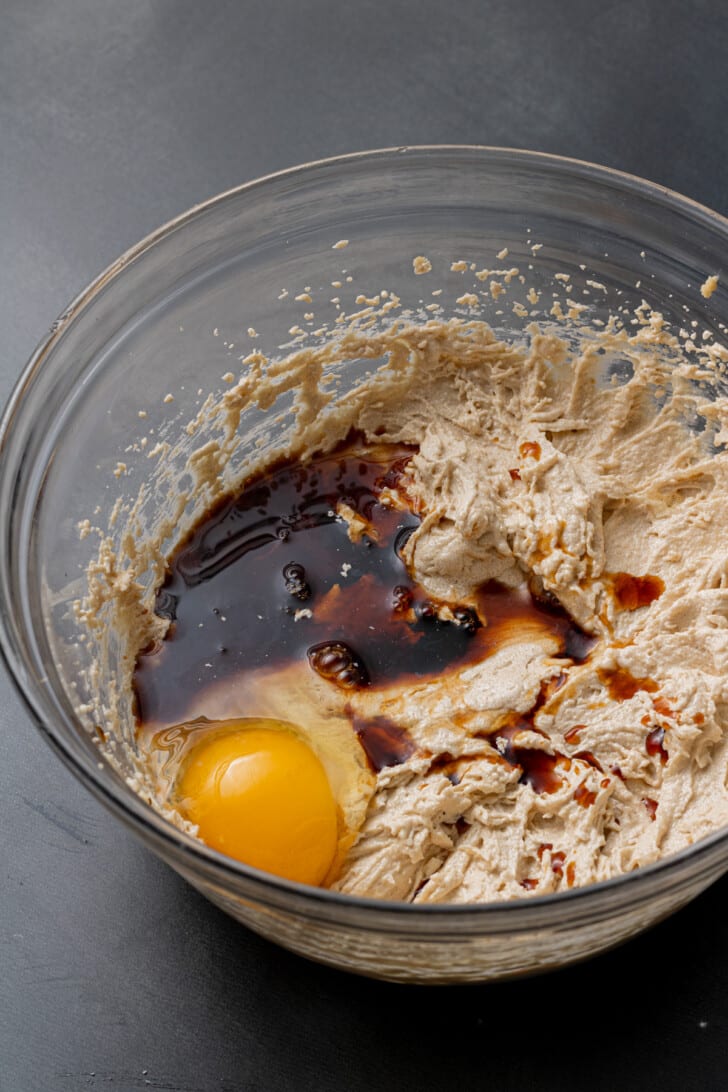Bowl with creamy batter, a whole egg, and dark liquid, possibly vanilla extract, blending together. Set on a dark surface, suggesting baking preparation.