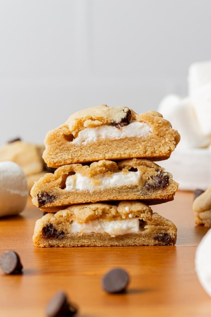 A stack of three cookies, halved to reveal gooey marshmallow centers. Chocolate chips are scattered on a wooden surface, creating a warm and tempting scene.