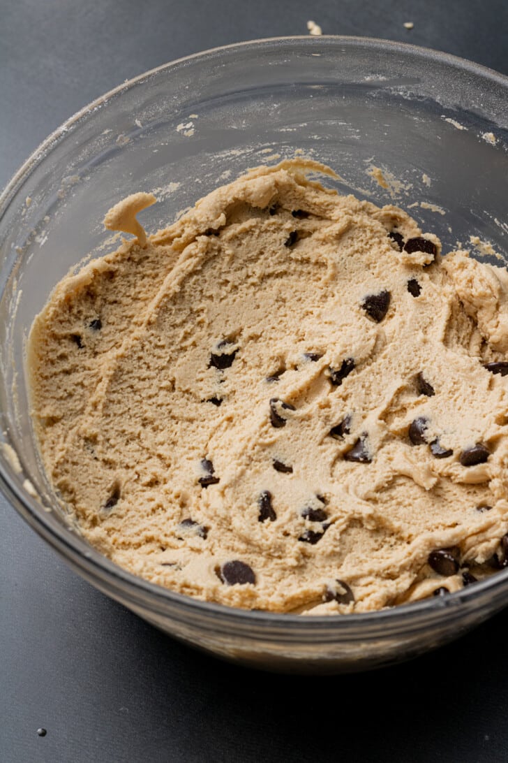 A clear glass bowl filled with creamy chocolate chip cookie dough sits on a dark countertop, evoking a sense of homemade comfort and anticipation.