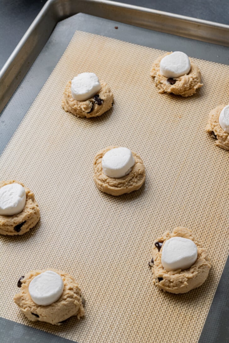 Cookie dough balls with chocolate chips, topped with marshmallows, are arranged on a baking sheet lined with parchment, ready for baking.