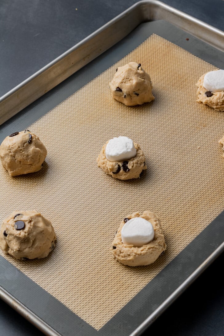 Six cookie dough balls with chocolate chips and marshmallow pieces are arranged on a baking sheet lined with a silicone mat, ready for baking.