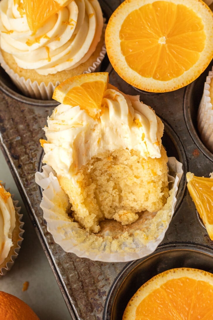 Orange cupcake with orange icing and a wedge of orange on top sits in a muffin tray. A bite is taken, revealing fluffy cake. Sliced orange halves nearby.