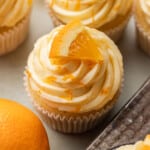 Orange cupcakes with swirled orange buttercream frosting topped with orange slices. The frosting is lightly sprinkled with orange zest, evoking a fresh, citrusy tone.