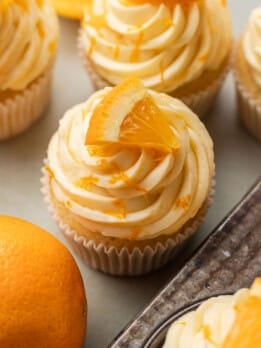 Orange cupcakes with swirled orange buttercream frosting topped with orange slices. The frosting is lightly sprinkled with orange zest, evoking a fresh, citrusy tone.