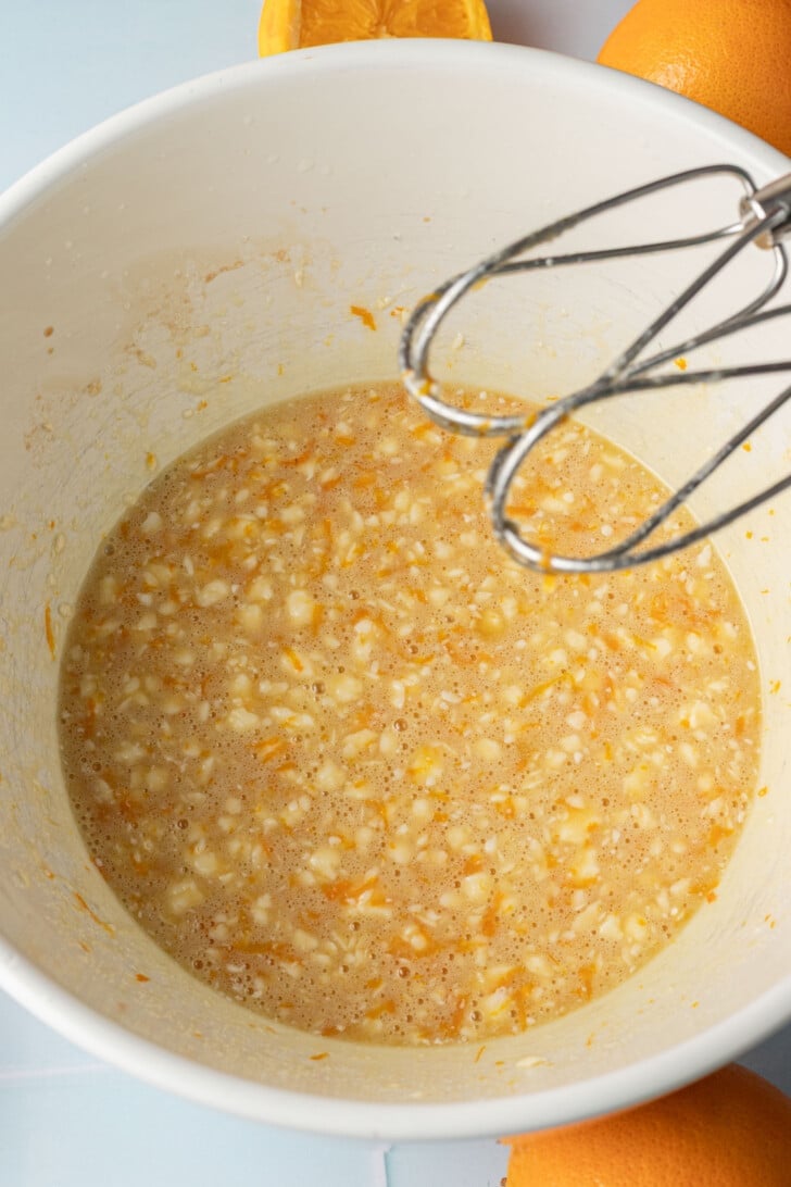 A mixing bowl containing an orange and beige batter with visible zest. A mixer is partially submerged, and fresh oranges are in the background.
