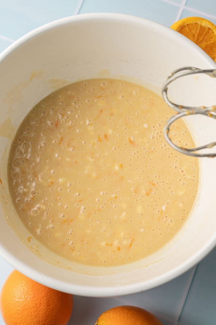A white bowl filled with a light orange batter and visible zest, next to an electric mixer. Whole and halved oranges are nearby on a tiled surface.