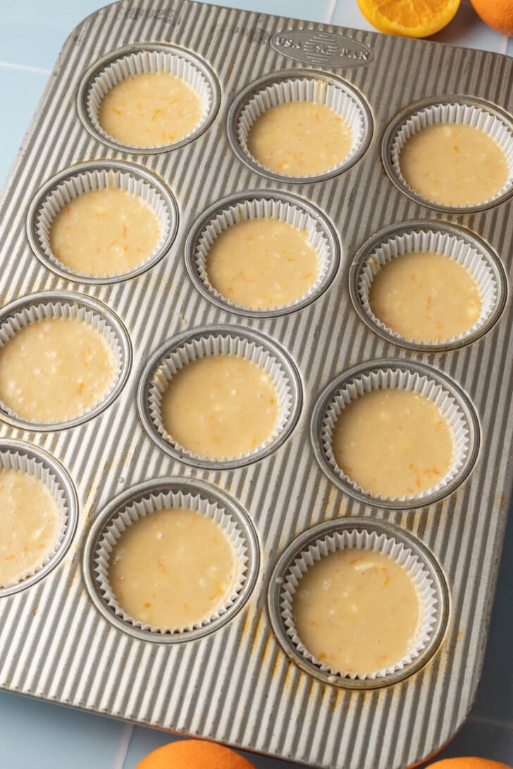 Muffin tin with 12 lined cups filled with orange-speckled batter. Halved oranges nearby, evoking a fresh, citrusy baking theme.