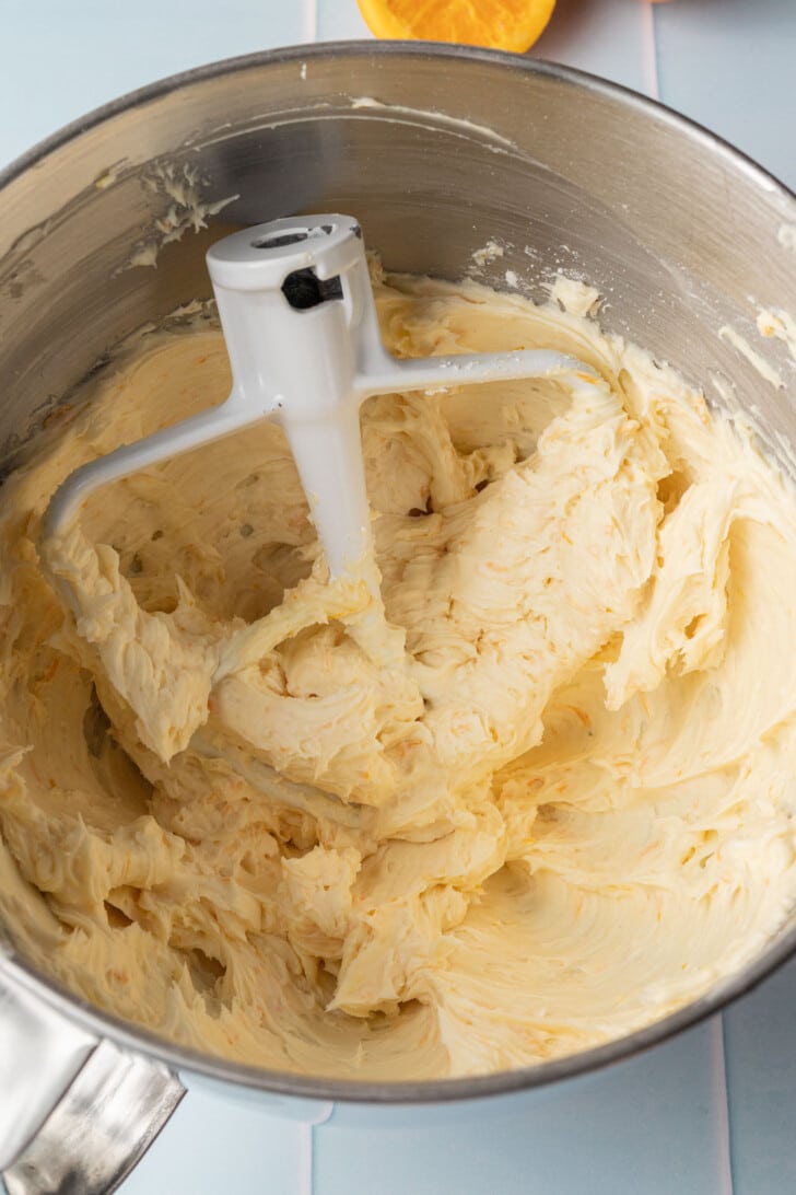 Close-up of orange buttercream mixture showing in a metal mixing bowl with a stand mixer paddle attachment. An orange slice is partially visible at the top.