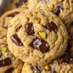 Close-up of stacked chocolate chip cookies with pistachios, topped with flaky sea salt. The cookies look warm and freshly baked, conveying a homemade feel.