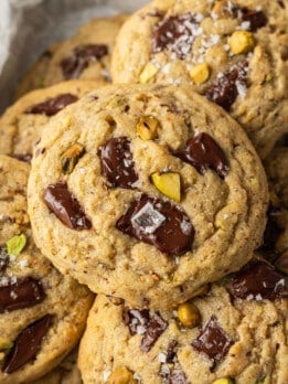Close-up of stacked chocolate chip cookies with pistachios, topped with flaky sea salt. The cookies look warm and freshly baked, conveying a homemade feel.
