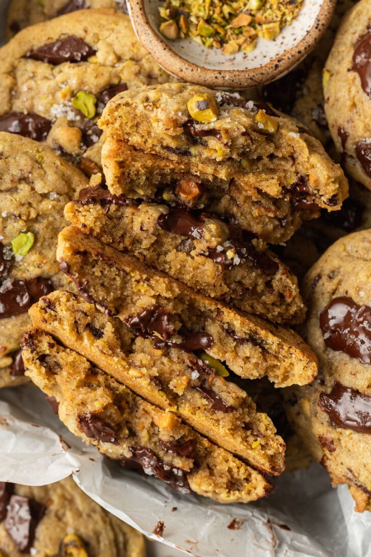 Close-up of freshly baked cookies with gooey chocolate chunks and sprinkled pistachios, some cookies are broken in half. Warm and inviting appearance.