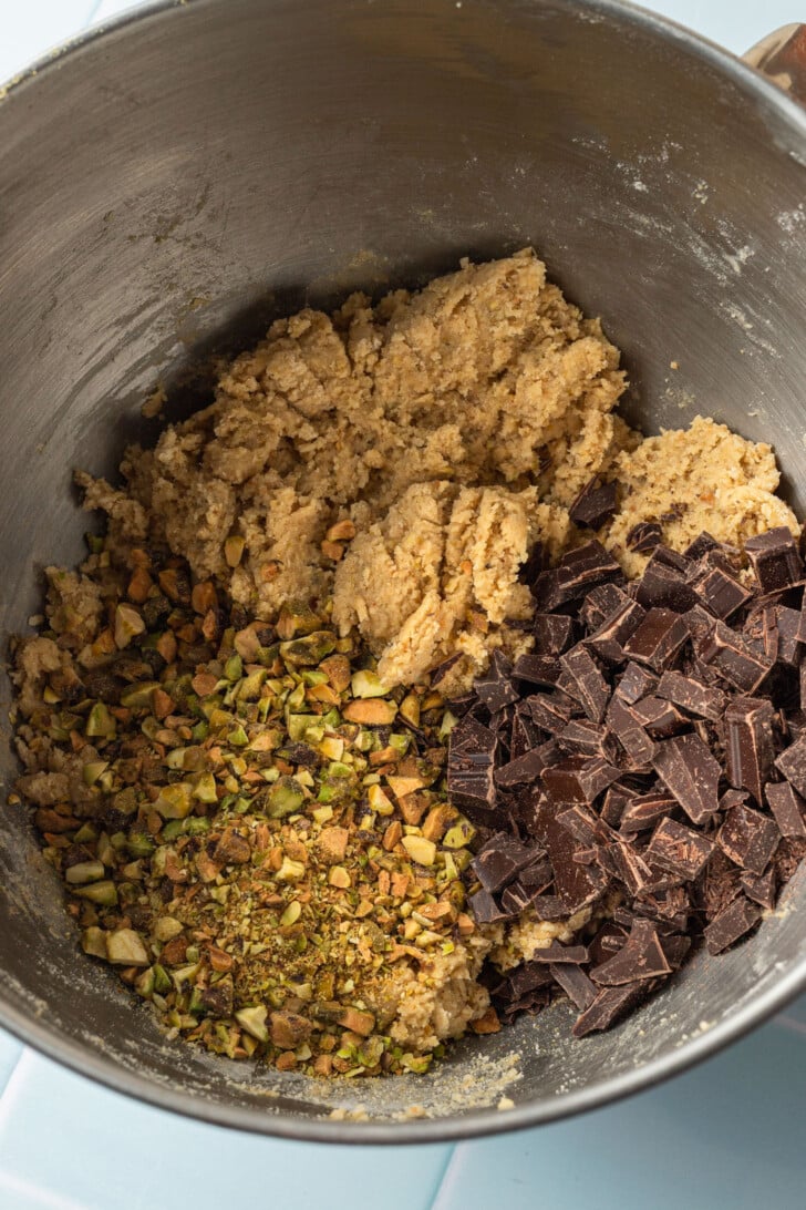 Bowl with cookie dough, chopped pistachios, and dark chocolate chunks ready for mixing. The mood is cozy and inviting, highlighting baking preparations.
