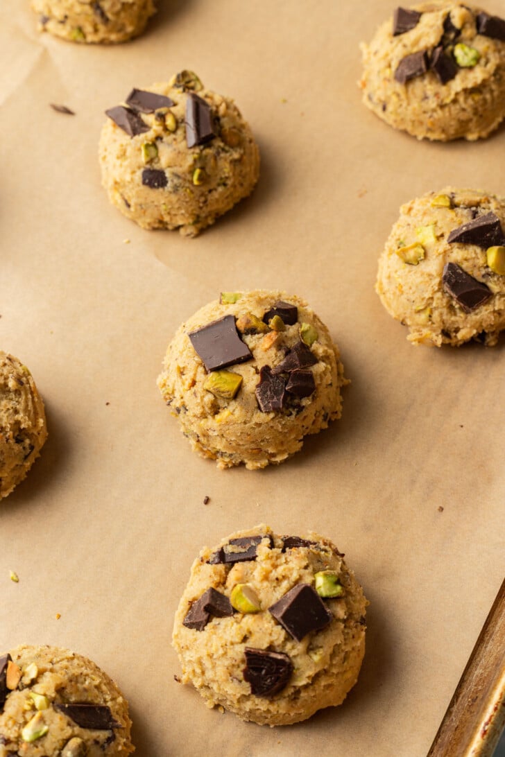 Scoops of cookie dough with dark chocolate chunks and green pistachios on a parchment-lined baking sheet, ready to bake. Inviting and homemade.