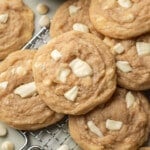 Close-up of freshly baked cookies with white chocolate chunks on a cooling rack. A wooden bowl with white chips adds a cozy, homemade feel.