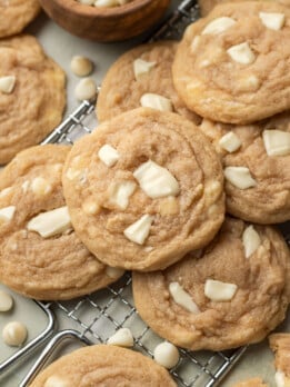 Close-up of freshly baked cookies with white chocolate chunks on a cooling rack. A wooden bowl with white chips adds a cozy, homemade feel.