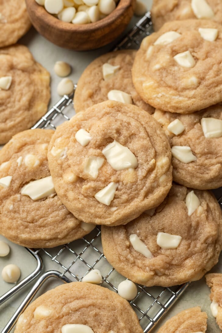 Close-up of freshly baked cookies with white chocolate chunks on a cooling rack. A wooden bowl with white chips adds a cozy, homemade feel.