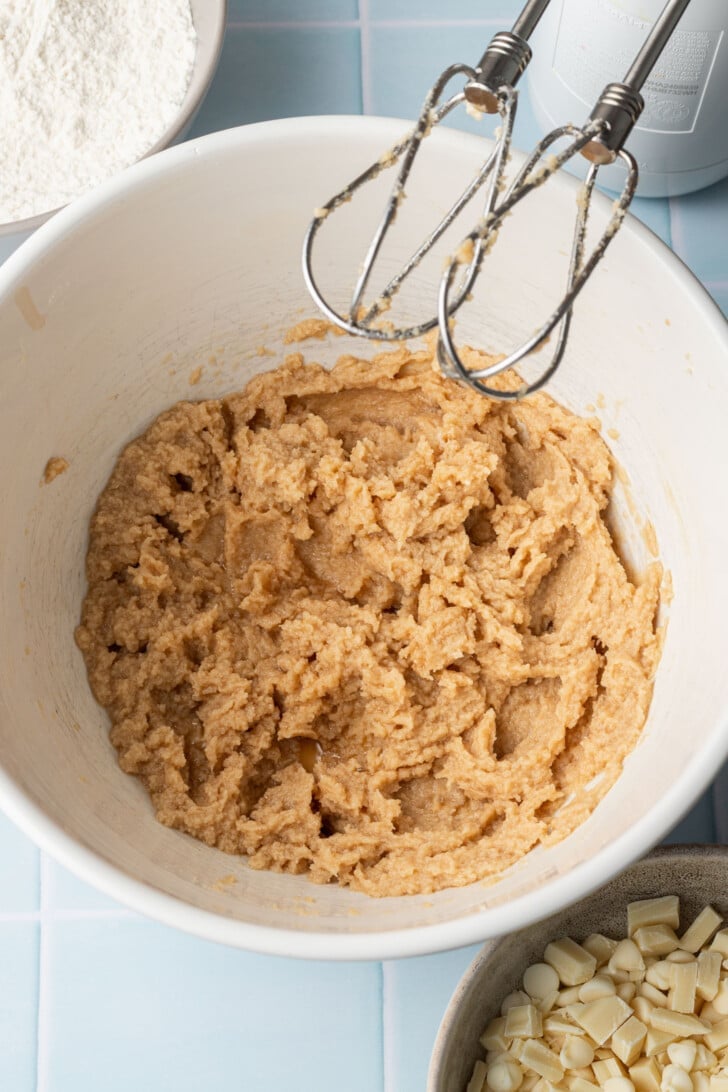 Mixing bowl with cookie dough and hand mixer on a light blue tile counter. Nearby are a bowl of flour and white chocolate chips, ready for baking.