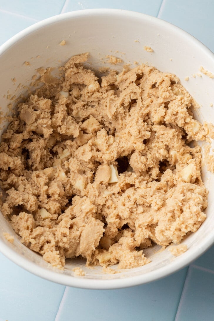 Cookie dough with white chocolate chunks in a cream-colored bowl on a light blue tiled surface, evoking a cozy, homemade baking vibe.