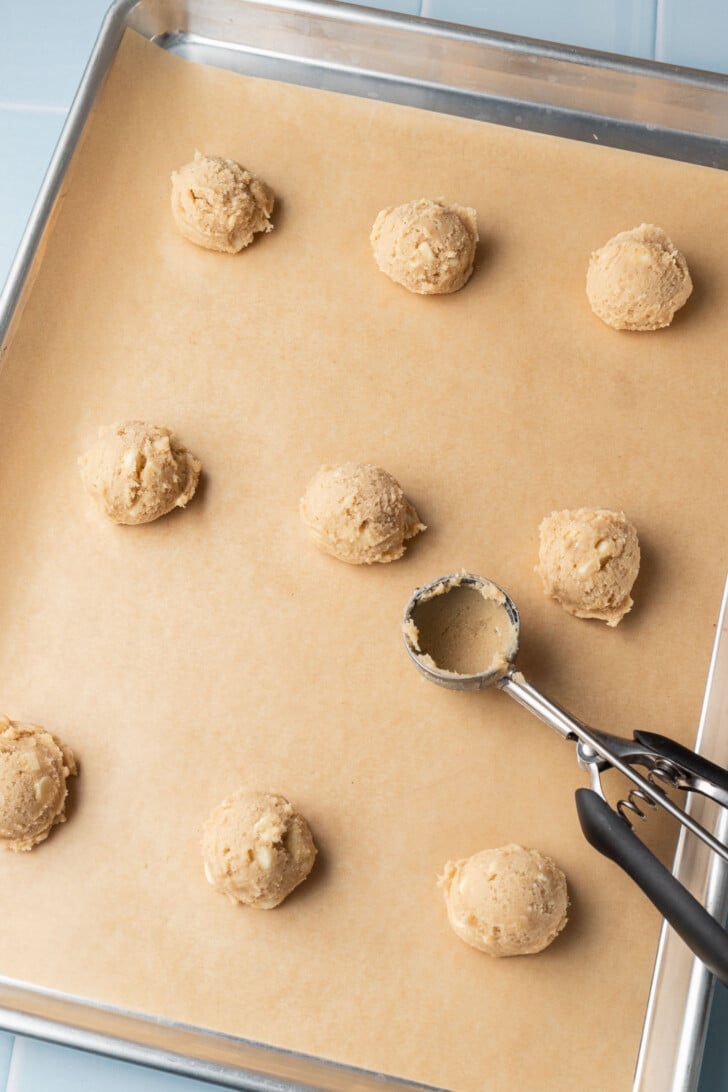Scoops of cookie dough spaced evenly on a parchment-lined baking sheet, with a cookie dough scoop resting beside them, ready for baking.