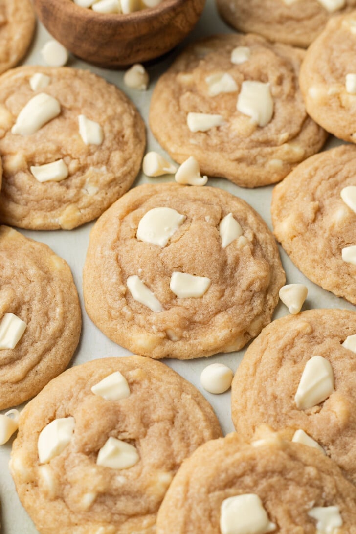 Close-up of freshly baked white chocolate chip cookies on a light surface, with a wooden bowl of chips in the background. The tone is cozy and inviting.
