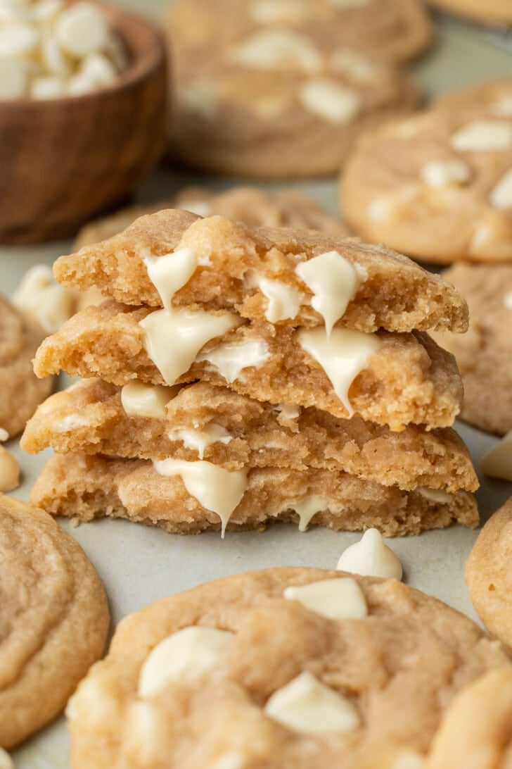 A stack of three thick, gooey white chocolate chip cookies, broken in half to reveal melty chocolate. Surrounding cookies are soft and golden brown.