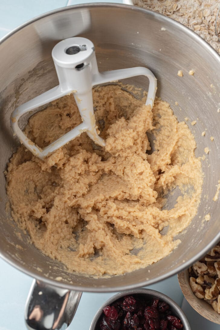 Close-up of a stand mixer bowl filled with creamy cookie dough. Nearby are small bowls of dried cranberries and walnuts, suggesting a baking scene.