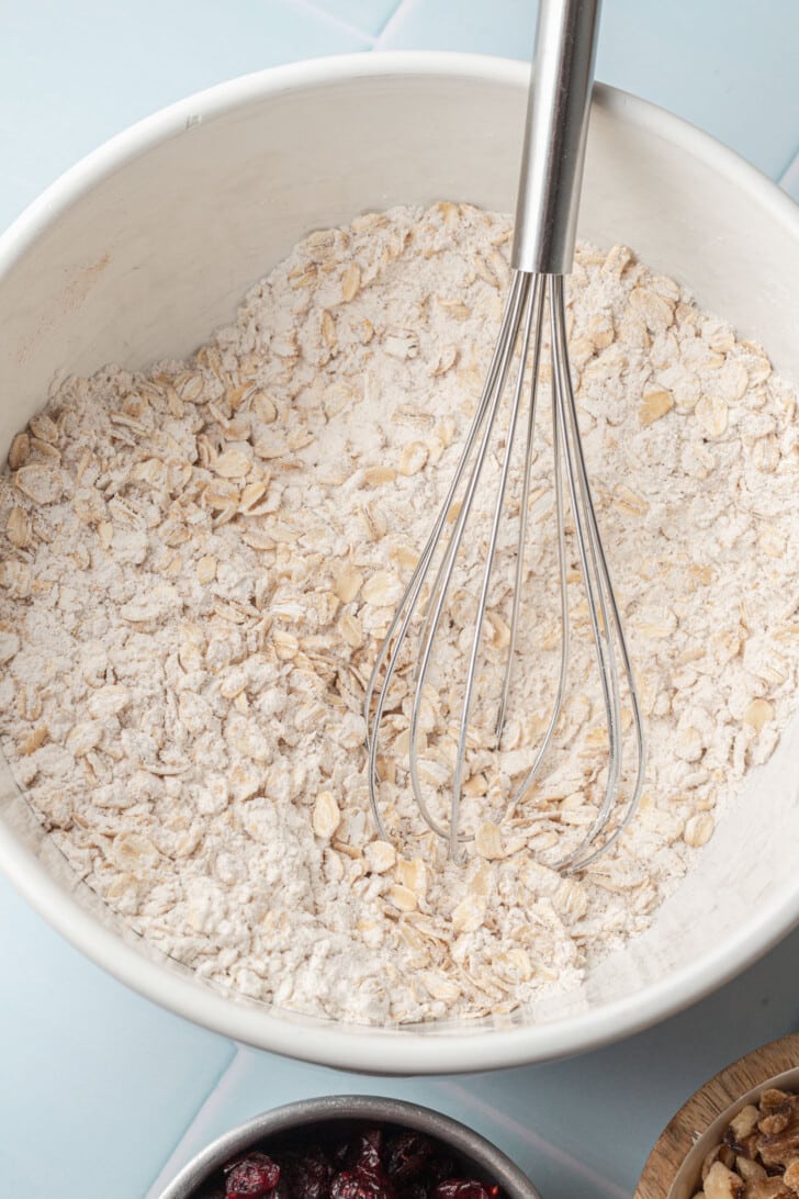A whisk resting in a white bowl filled with oats and flour on a light blue surface. Below the bowl, a small dish of dried cranberries is partially visible.